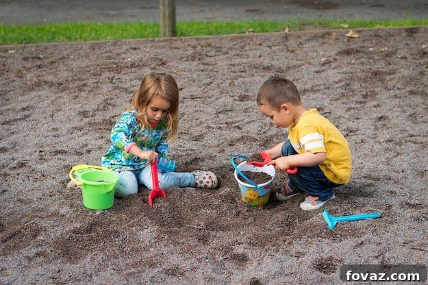 A large wooden sandbox with a hinged wooden cover that folds out into two benches, one on each side, revealing the sandpit inside. The sandbox is situated on a grassy lawn.