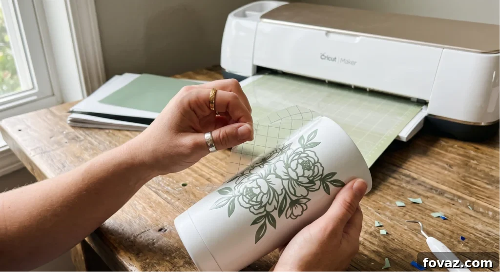Woman peeling transfer tape off a finished floral vinyl decal on a white tumbler, with a Cricut Maker visible in the background — one of the most popular cricut projects to sell from home.